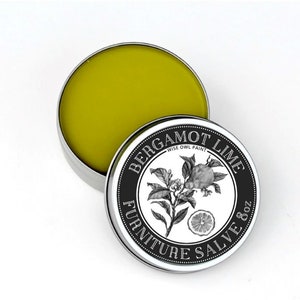 Può includere: Un contenitore rotondo di latta con un'etichetta in bianco e nero che recita "Bergamot Lime Furniture Salve 8 oz - Wise Owl Paint". L'etichetta presenta un'illustrazione in bianco e nero di un albero di bergamotto con frutti e fiori. La latta è aperta e il contenitore è pieno di una pomata gialla.