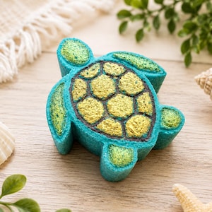 May include: A sea turtle shaped bath bomb with a bright blue body and a textured shell featuring gold and green accents. The shell is composed of several raised, rounded segments outlined in black, while the flippers and head have a shimmering finish.