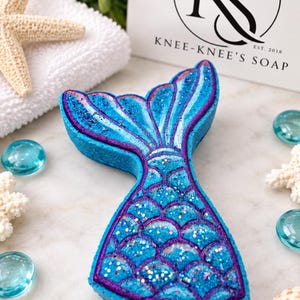 May include: A mermaid tail-shaped bath bomb in shades of blue and purple with a glittery finish. The surface features a molded scale pattern. A white card with the black text KS KNEE-KNEE'S SOAP EST. 2018 is positioned behind the bath bomb.