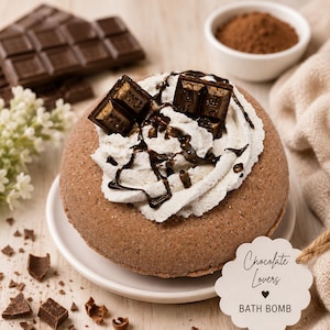 May include: A round, brown, textured bath bomb topped with white whipped cream, dark chocolate drizzle, and two chocolate squares. A scalloped white tag attached with twine reads "Chocolate Lovers BATH BOMB" in black script and print text.