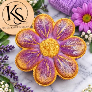 May include: A flower-shaped bath bomb with purple petals featuring gold-dusted edges and a textured gold center. A circular wooden sign nearby displays the text KS KNEE-KNEE'S SOAP EST. 2018. The bath bomb is surrounded by purple lavender and a pink flower.
