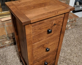 May include: A wooden nightstand with three drawers. The nightstand is stained a warm brown color and has black square knobs. The top of the nightstand has a slightly overhanging edge. The nightstand is approximately 28 inches tall.