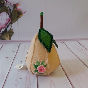 May include: A beige felt pear with a green felt leaf and pink embroidered flowers. The pear has a brown stem with a green jewel on top.