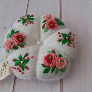 May include: White pincushion with a flower design embroidered in pink and green thread. The pincushion is shaped like a flower with five petals.