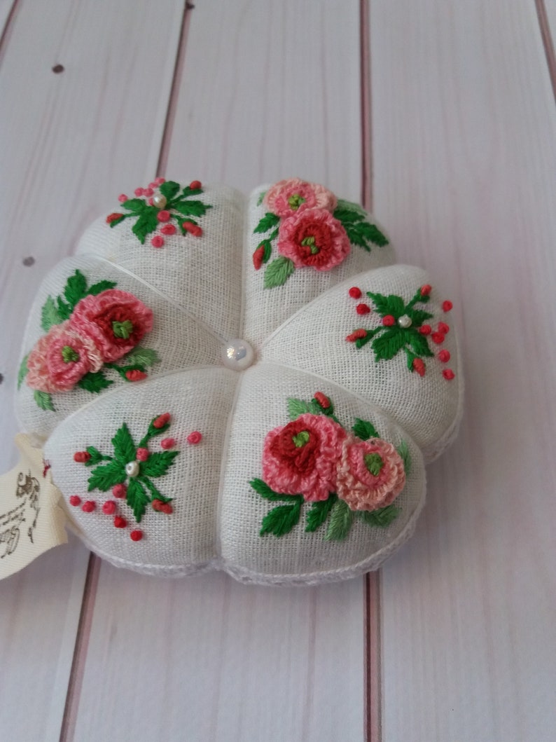 May include: White pincushion with a flower design embroidered in pink and green thread. The pincushion is shaped like a flower with five petals.