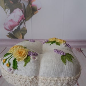 May include: A white, round pincushion with embroidered yellow roses, purple lavender, and green leaves. The pincushion has a cream-colored crocheted trim and is displayed on a white pedestal. The background features a floral print.