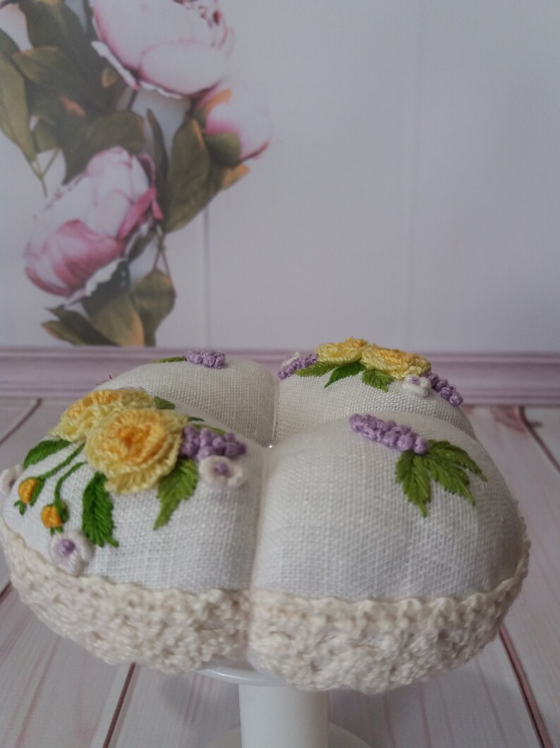 May include: A white, round pincushion with embroidered yellow roses, purple lavender, and green leaves. The pincushion has a cream-colored crocheted trim and is displayed on a white pedestal. The background features a floral print.