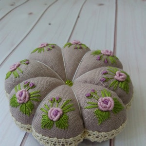 May include: A round, beige pincushion with a lace trim. The pincushion is decorated with embroidered pink flowers and green leaves.