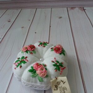 May include: White linen pincushion with pink and green embroidered flowers. The pincushion has a pearl button in the center and a white lace trim. A small tag with the text "Hand Made" is attached to the pincushion.