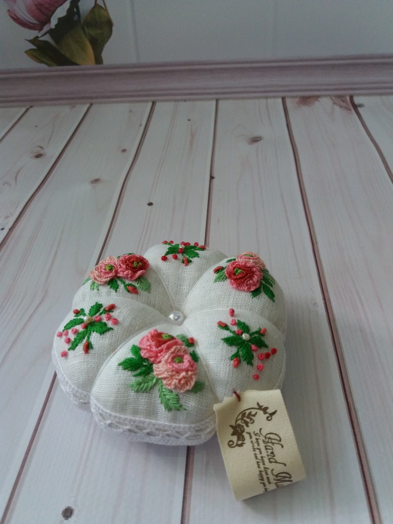May include: White linen pincushion with pink and green embroidered flowers. The pincushion has a pearl button in the center and a white lace trim. A small tag with the text "Hand Made" is attached to the pincushion.