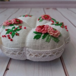 May include: A white heart-shaped pincushion with a lace trim and embroidered pink flowers and green leaves.