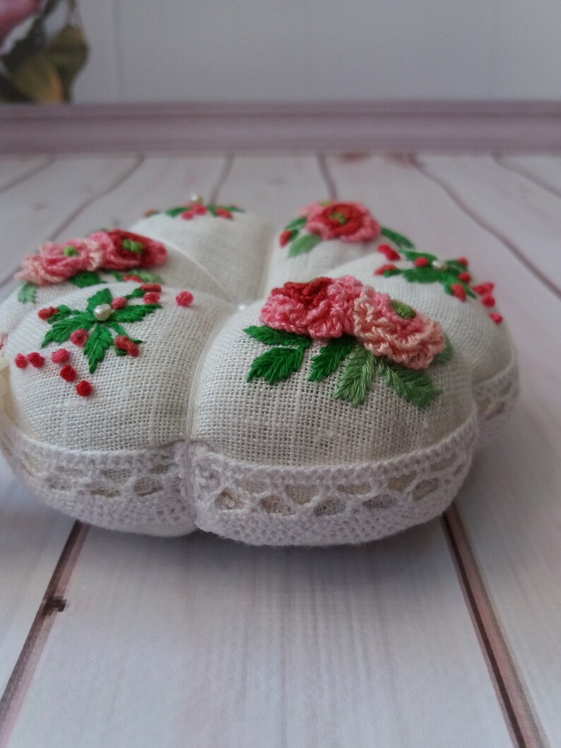 May include: A white heart-shaped pincushion with a lace trim and embroidered pink flowers and green leaves.