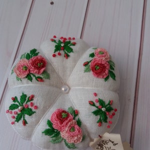 May include: A white fabric pincushion with a pearl button in the center. The pincushion is decorated with pink and red embroidered flowers and green leaves.