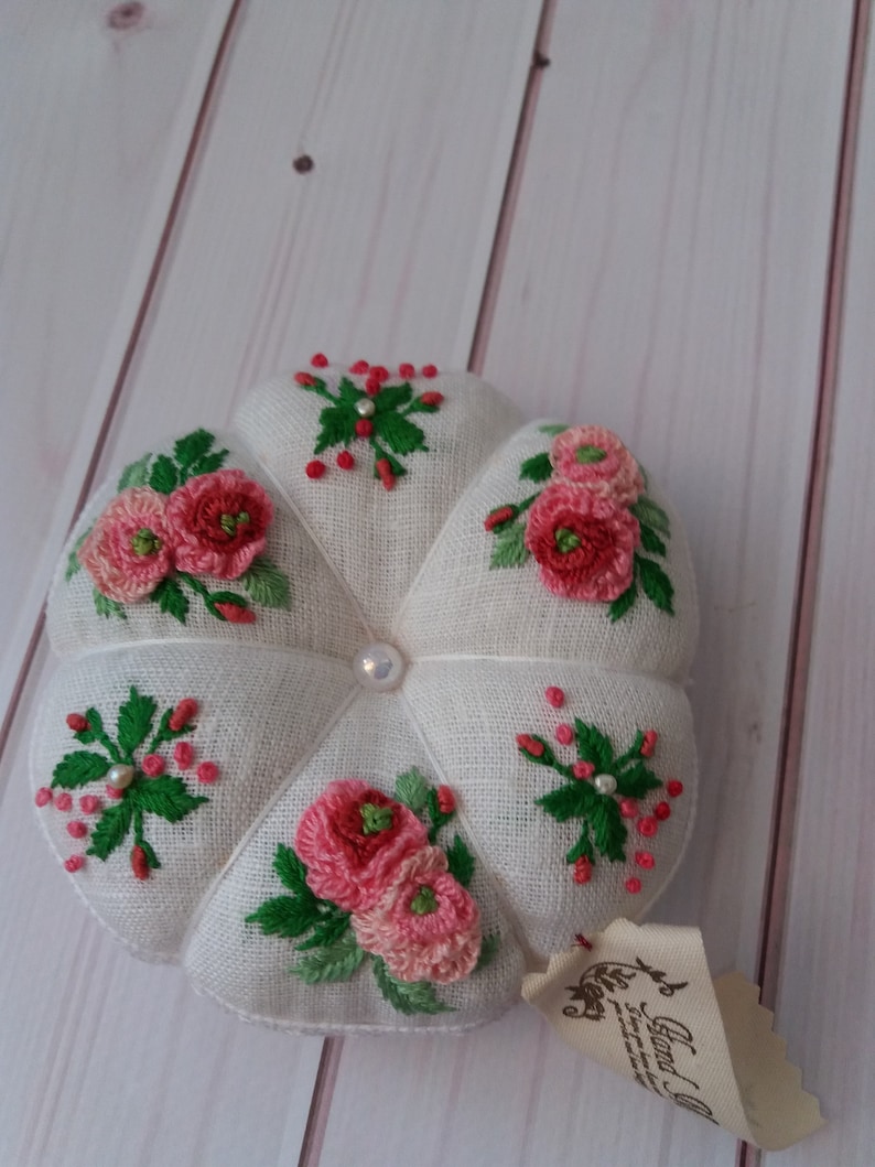 May include: A white fabric pincushion with a pearl button in the center. The pincushion is decorated with pink and red embroidered flowers and green leaves.