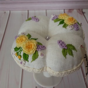 May include: A white, four-sectioned pincushion with embroidered yellow roses and purple lilacs. The pincushion has a crocheted edge and sits on a white pedestal. A small tag with the word "handmade" is attached.