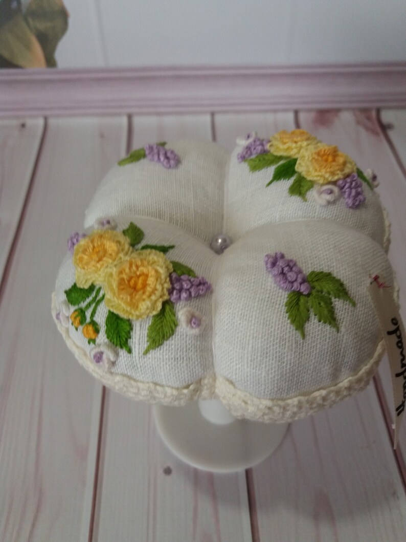 May include: A white, four-sectioned pincushion with embroidered yellow roses and purple lilacs. The pincushion has a crocheted edge and sits on a white pedestal. A small tag with the word "handmade" is attached.
