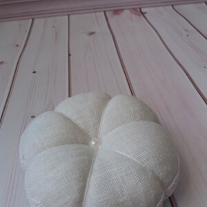 May include: A white, round, fabric ring pillow with a pearl button in the center. The pillow has a scalloped edge and is made of a textured linen fabric.