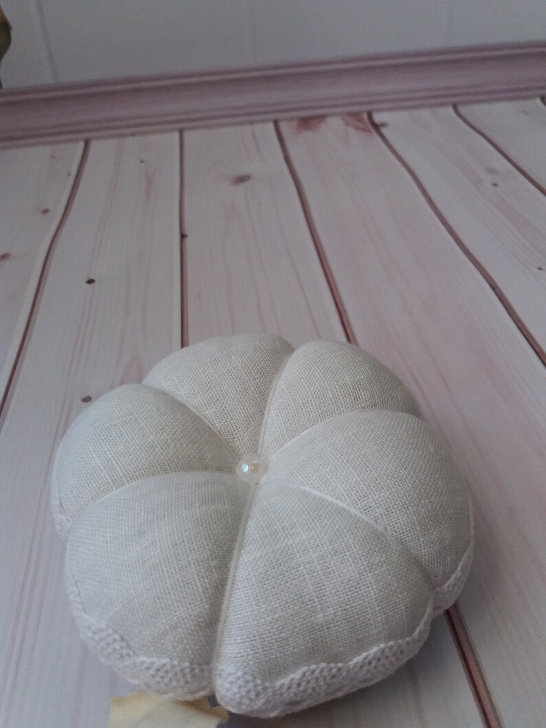 May include: A white, round, fabric ring pillow with a pearl button in the center. The pillow has a scalloped edge and is made of a textured linen fabric.
