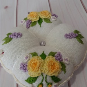 May include: A white, four-sectioned pincushion with embroidered floral designs. The pincushion features yellow roses, purple clusters, and green leaves. A pearl bead is centered on the top. The edges are trimmed with a cream-colored crochet.