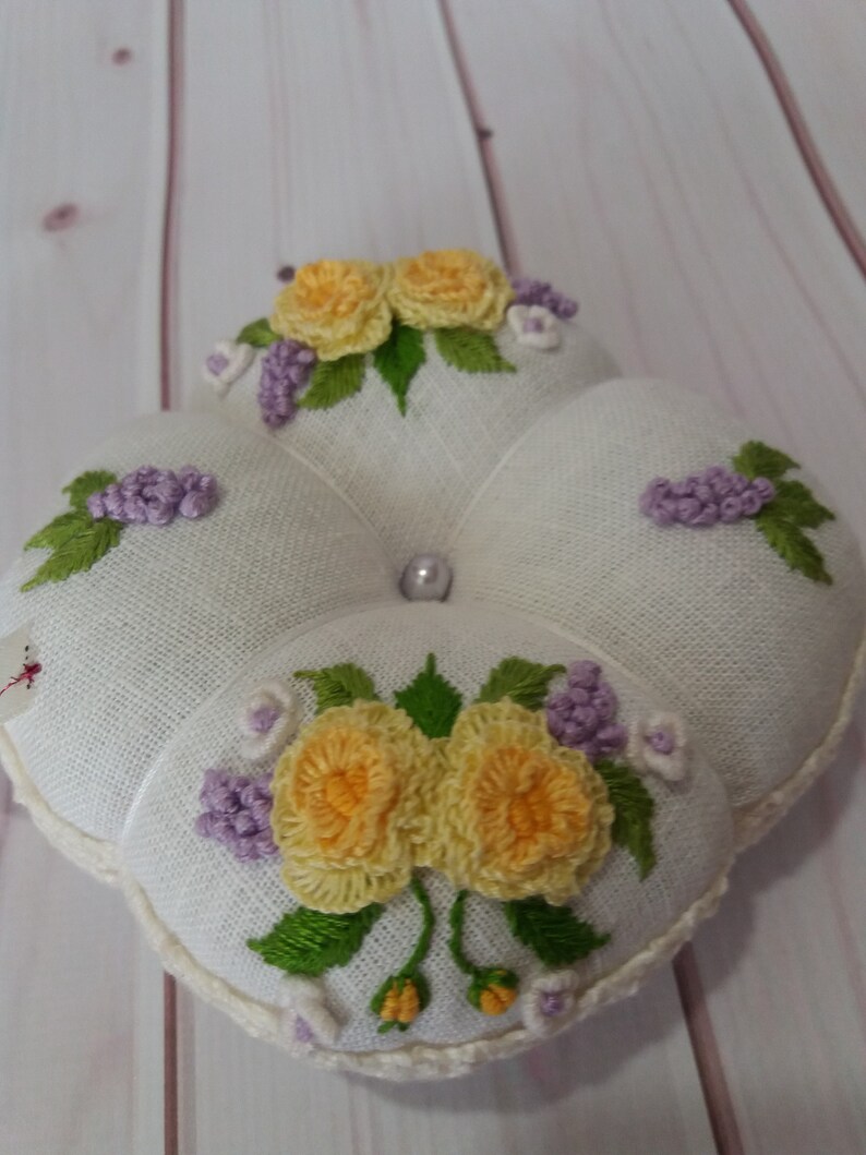May include: A white, four-sectioned pincushion with embroidered floral designs. The pincushion features yellow roses, purple clusters, and green leaves. A pearl bead is centered on the top. The edges are trimmed with a cream-colored crochet.