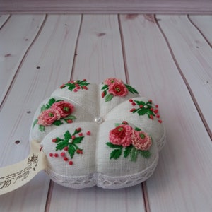 May include: White linen pincushion with a flower design embroidered in pink and green. The pincushion is shaped like a flower and has a pearl button in the center.
