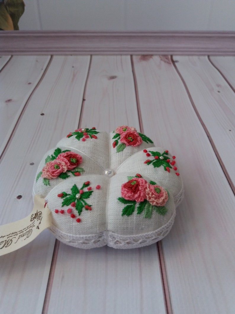 May include: White linen pincushion with a flower design embroidered in pink and green. The pincushion is shaped like a flower and has a pearl button in the center.