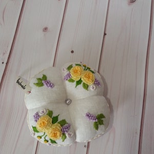 May include: A white, four-sectioned pincushion with embroidered yellow roses and purple flowers. A small tag reads "handmade". A pearl bead is in the center. The pincushion is on a light wood surface.