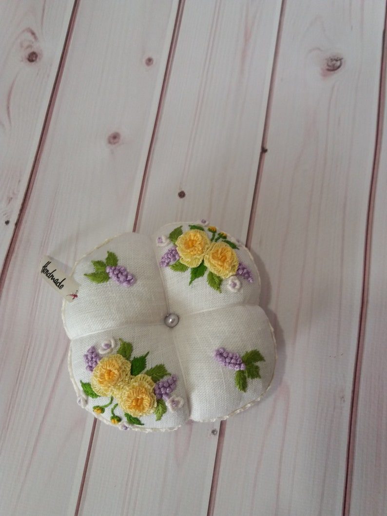 May include: A white, four-sectioned pincushion with embroidered yellow roses and purple flowers. A small tag reads "handmade". A pearl bead is in the center. The pincushion is on a light wood surface.