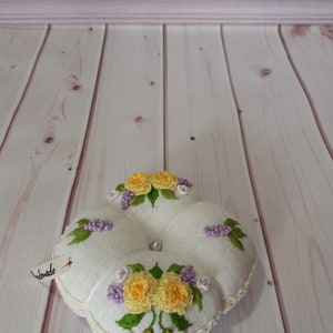 May include: A handmade, white pin cushion with embroidered yellow roses and purple flowers. The cushion has a crocheted edge and a pearl bead center. A small tag reads "handmade".