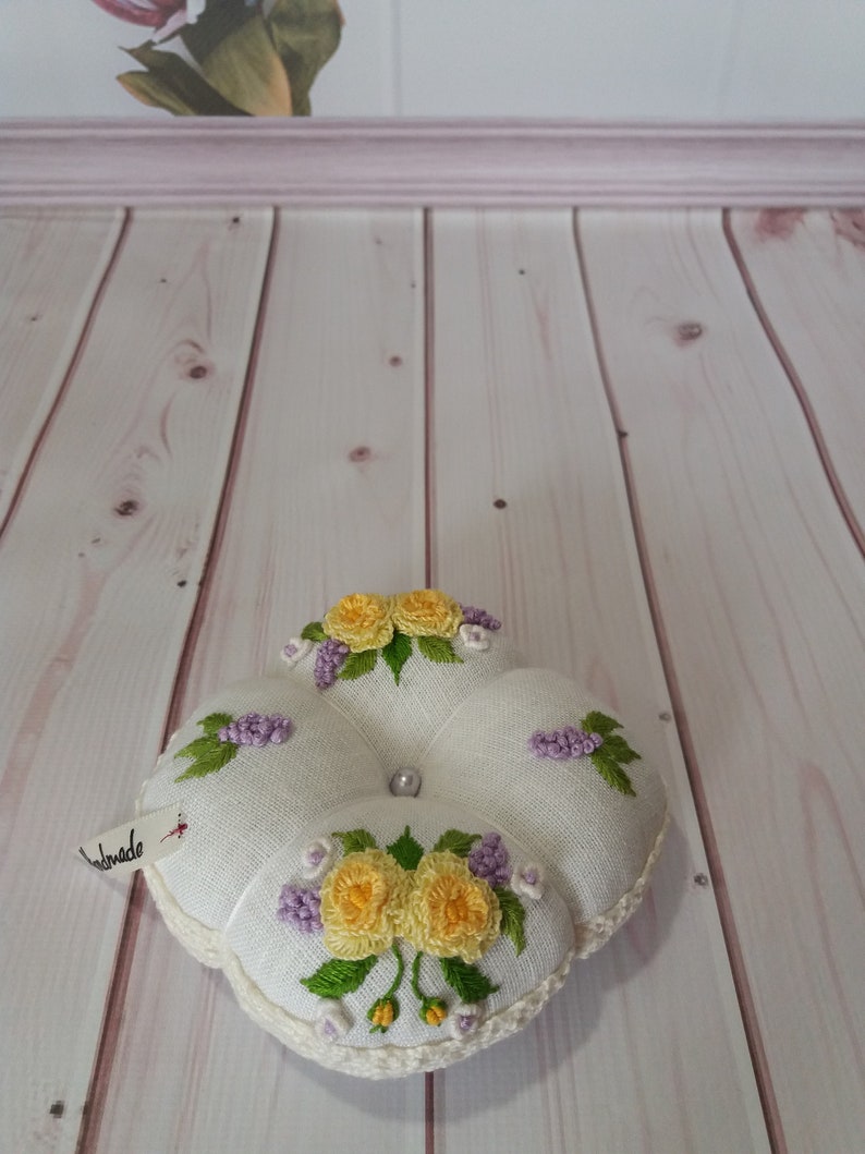 May include: A handmade, white pin cushion with embroidered yellow roses and purple flowers. The cushion has a crocheted edge and a pearl bead center. A small tag reads "handmade".