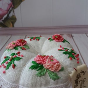 May include: White linen pincushion with a heart shape and embroidered pink flowers and green leaves. The pincushion has a white lace trim and is sitting on a white pedestal. The pincushion has a tag that says "Hand Made".