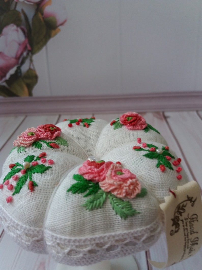 May include: White linen pincushion with a heart shape and embroidered pink flowers and green leaves. The pincushion has a white lace trim and is sitting on a white pedestal. The pincushion has a tag that says "Hand Made".