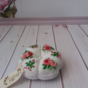 May include: A white pincushion with a lace trim and embroidered pink flowers. The pincushion is sitting on a white wooden surface.