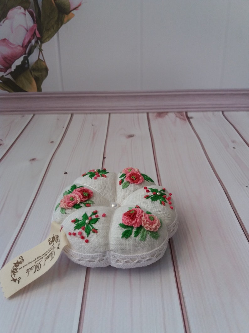May include: White pincushion with a lace trim and embroidered pink flowers and green leaves. The pincushion is sitting on a white wooden surface.