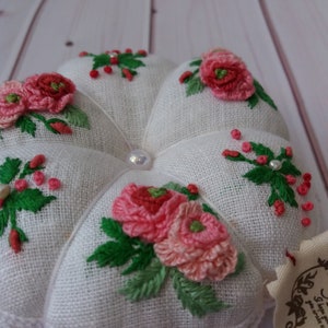 May include: White linen pincushion with a heart shape and embroidered pink and red flowers with green leaves. The pincushion has a white lace trim and a pearl button in the center.