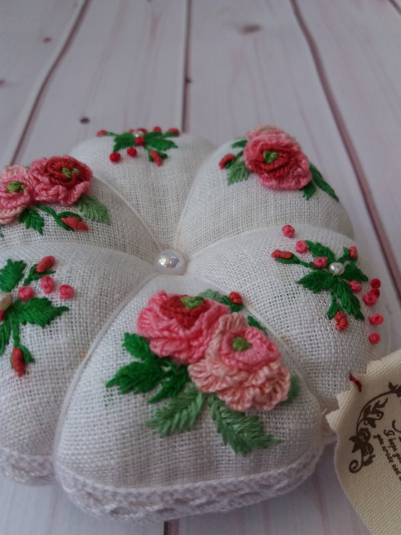 May include: White linen pincushion with a heart shape and embroidered pink and red flowers with green leaves. The pincushion has a white lace trim and a pearl button in the center.