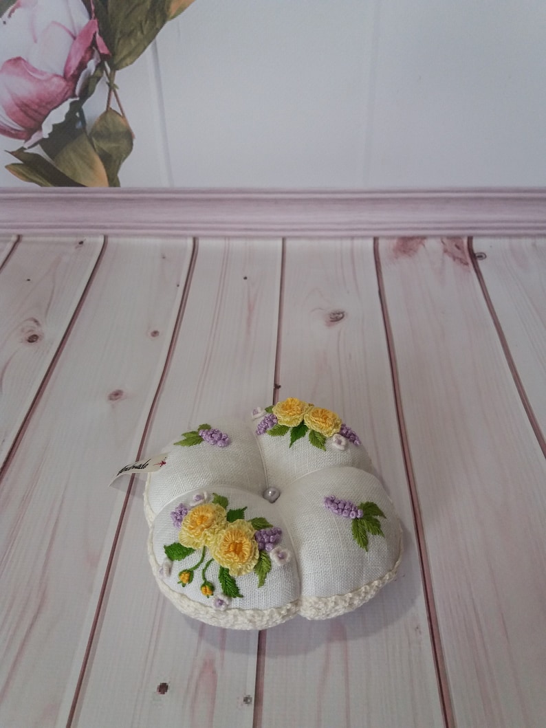 May include: A white, flower-shaped pincushion with yellow and purple floral embroidery. The pincushion has a crocheted edge and a pearl bead in the center. The background features a floral painting and light wood paneling.