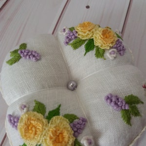 May include: A white, four-sectioned pin cushion with embroidered floral designs. The cushion features yellow roses, purple clusters, and green leaves. A pearl bead is centered. The edges are trimmed with a delicate, cream-colored lace.