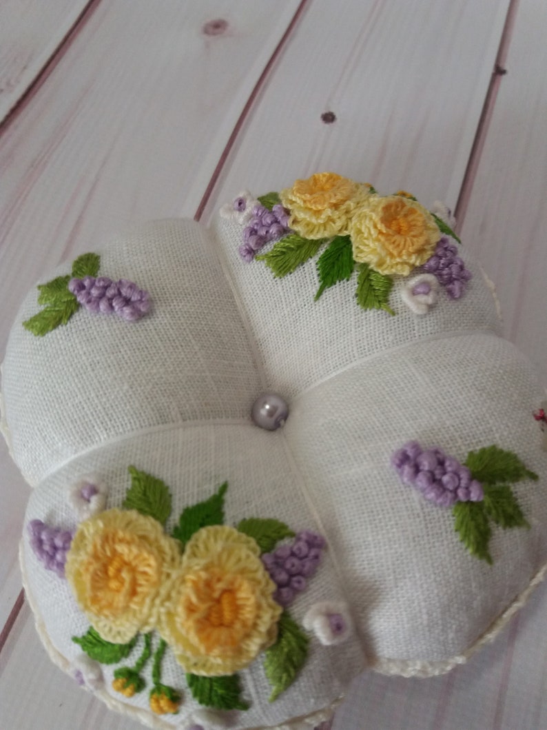 May include: A white, four-sectioned pin cushion with embroidered floral designs. The cushion features yellow roses, purple clusters, and green leaves. A pearl bead is centered. The edges are trimmed with a delicate, cream-colored lace.
