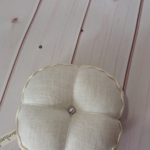 May include: A cream-colored, four-lobed pincushion with a pearl bead center. The pincushion is made of a textured fabric and has a decorative, cream-colored trim around the edges. A small tag is attached to the side.