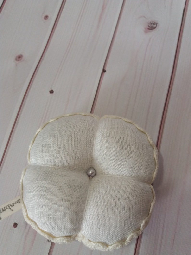 May include: A cream-colored, four-lobed pincushion with a pearl bead center. The pincushion is made of a textured fabric and has a decorative, cream-colored trim around the edges. A small tag is attached to the side.