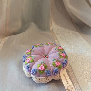 May include: A round, lavender-colored pincushion featuring embroidered pink roses, green leaves, and blue floral accents. The cushion has a tufted design with a central bead detail and a cream-colored lace trim. A small tag attached reads Hand Made.