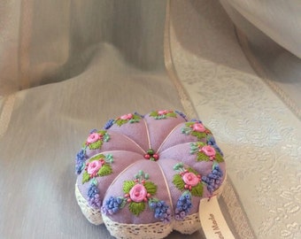 May include: A round, lavender-colored pincushion featuring embroidered pink roses, green leaves, and blue floral accents. The cushion has a tufted design with a central bead detail and a cream-colored lace trim. A small tag attached reads Hand Made.