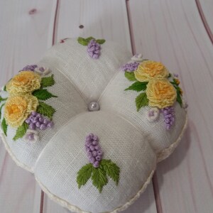 May include: A white, four-lobed pincushion with embroidered yellow roses, purple grapes, and green leaves. A pearl bead is centered on the top. The edges are trimmed with a cream-colored, crocheted border.