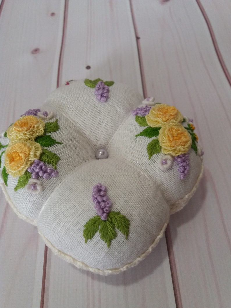 May include: A white, four-lobed pincushion with embroidered yellow roses, purple grapes, and green leaves. A pearl bead is centered on the top. The edges are trimmed with a cream-colored, crocheted border.