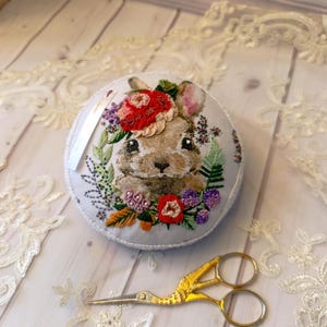 May include: A round, white pincushion with an embroidered bunny design surrounded by colorful flowers and greenery. Gold-colored scissors with a bird-like design are next to the pincushion. The pincushion has a white tag attached.