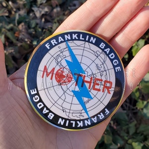 May include: A black and white button with a gold rim. The button features a world map with a blue lightning bolt and the text "MOTHER" in red letters. The text "FRANKLIN BADGE" is written around the edge of the button.