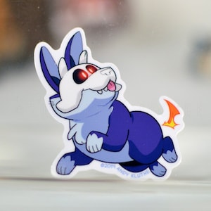 May include: A sticker featuring a cartoon creature with a white skull-like face, blue fur, and a dark purple body. It has red eyes and a small, orange flame-like tail. The sticker is on a clear surface.