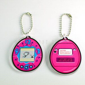 May include: Pink plastic keychain shaped like a Tamagotchi, with a digital pet on the screen and a clock face around the edge. The back of the keychain has a warning label about the battery.