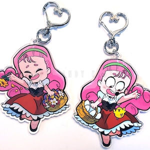 May include: Two clear acrylic keychains featuring a cartoon girl with pink hair, a red dress, and a basket of flowers or eggs. Each keychain has a silver heart-shaped clasp. The design is outlined in black.